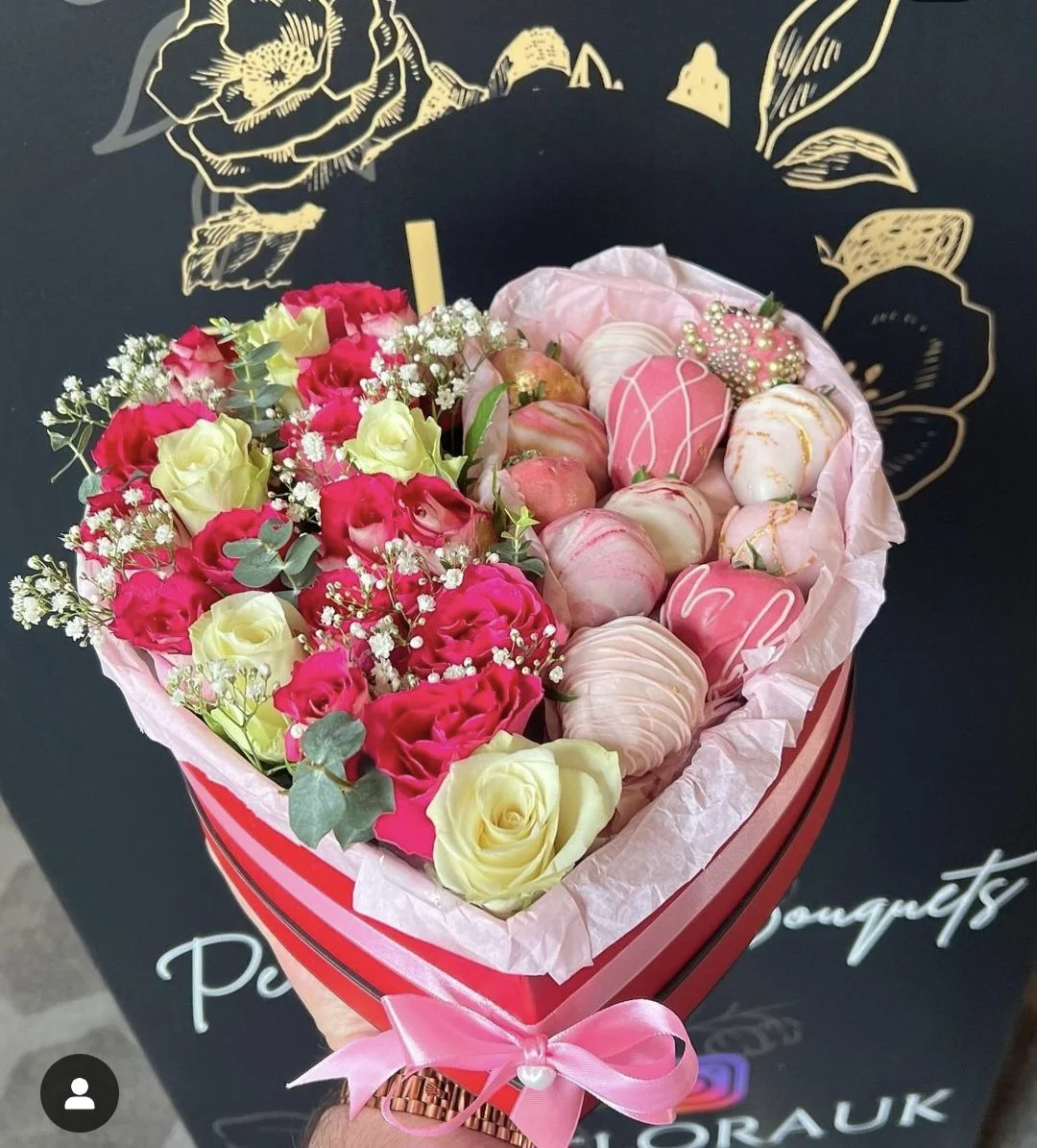 A heart-shaped bouquet of flowers and decorated cookies. The bouquet includes pink and white roses, baby's breath, and green foliage, wrapped in pink tissue paper with a pink ribbon. The cookies are pink and white, decorated with swirl and drip designs. The background is a black surface with gold floral illustrations and white text.