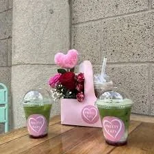 Two green drinks with straws in clear cups, pink heart-shaped plush, pink flowers in a pink box with a heart label, and a small green chair in the background.