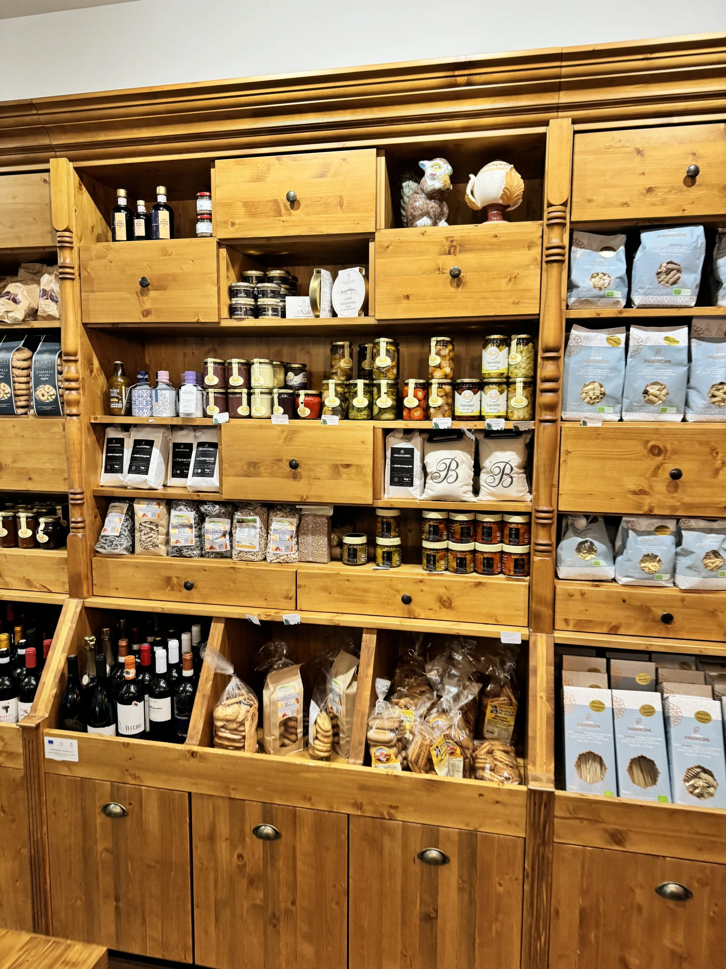 Wooden shelves stocked with jars of preserves, bags of pasta, and bottles of wine and olive oil in a grocery store