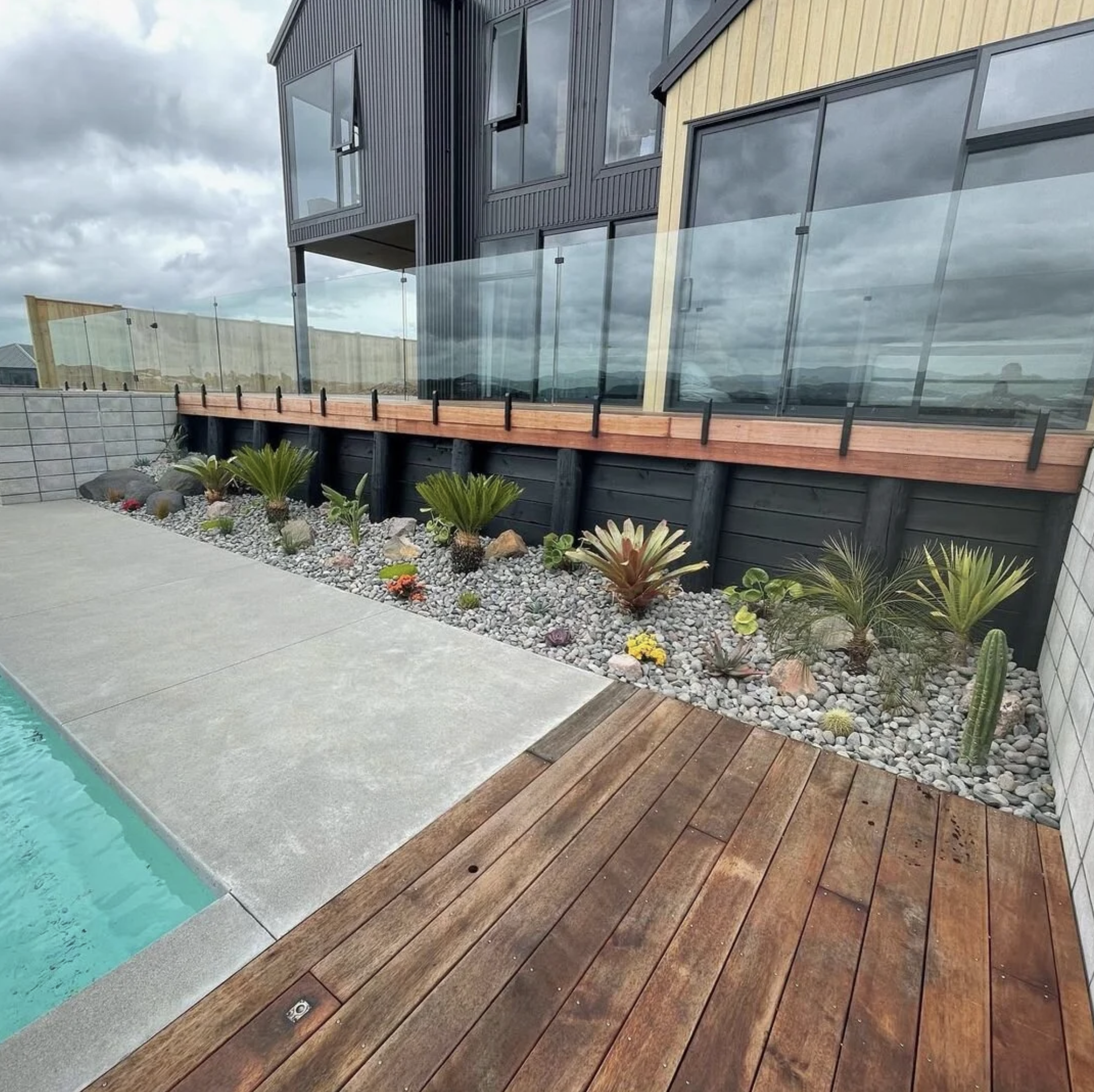 Exterior view of a modern house with black and beige siding, large windows, and a glass balcony, with a landscaped yard and cloudy sky in the background.