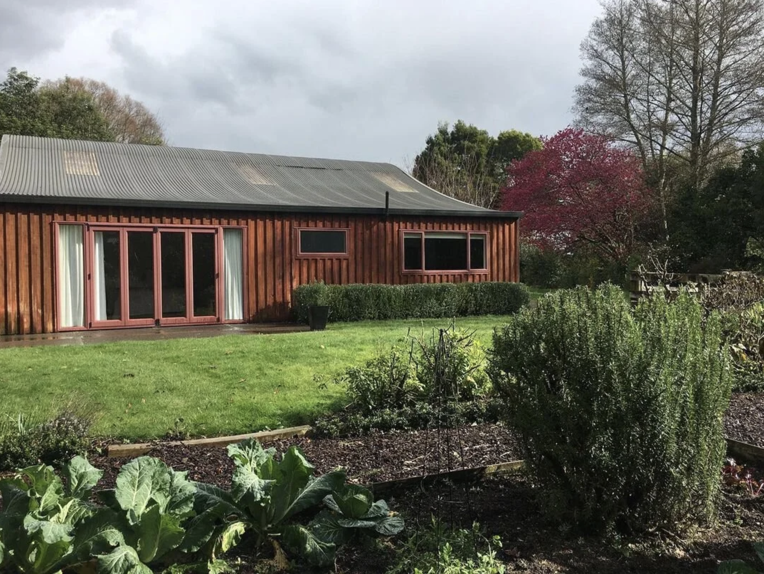 A suburban backyard with a wooden house, a garden bed, green grass, trees with autumn foliage, and a cloudy sky.