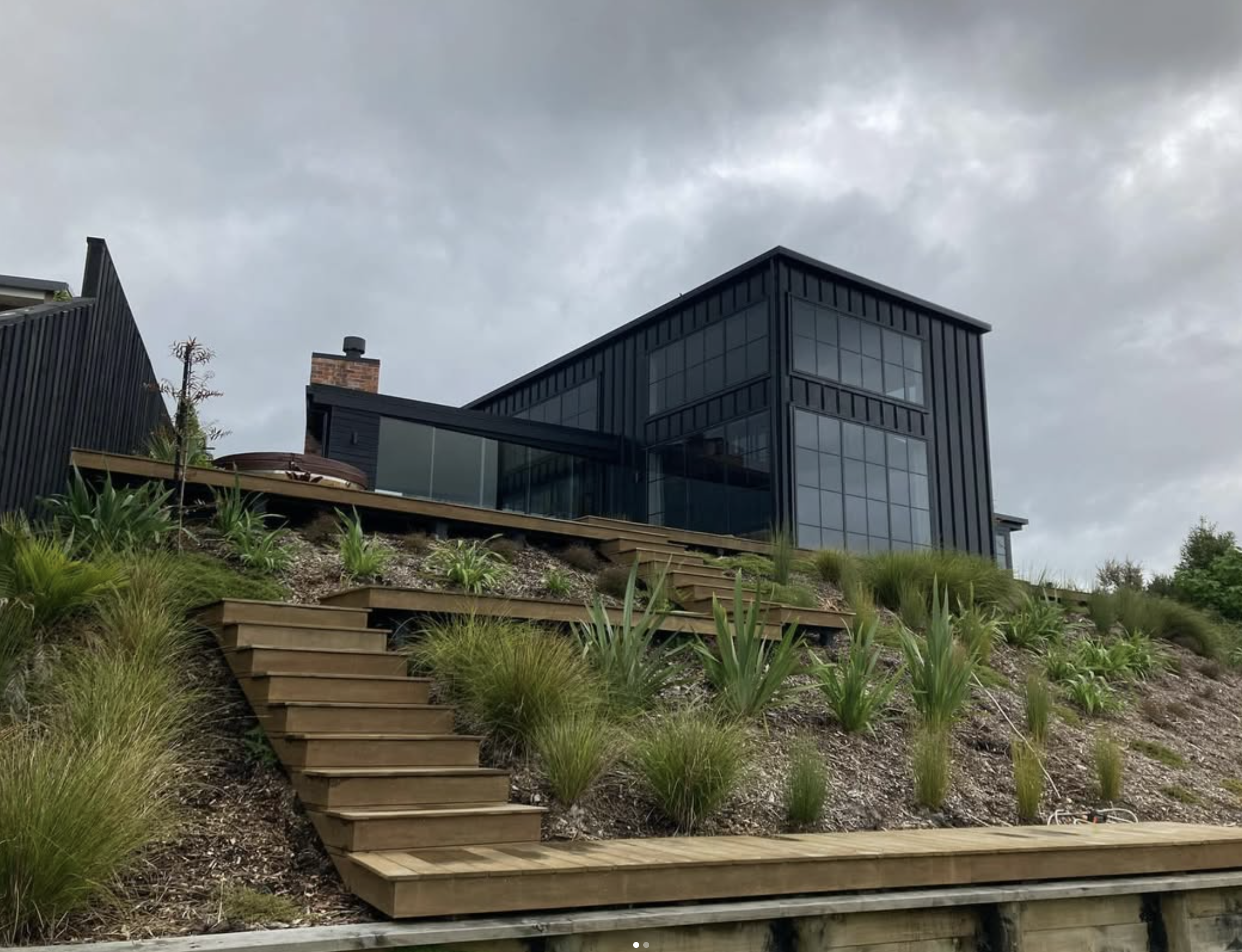 Modern black building with large glass windows and a chimney, seen from a garden with wooden stairs and green plants on a cloudy day.