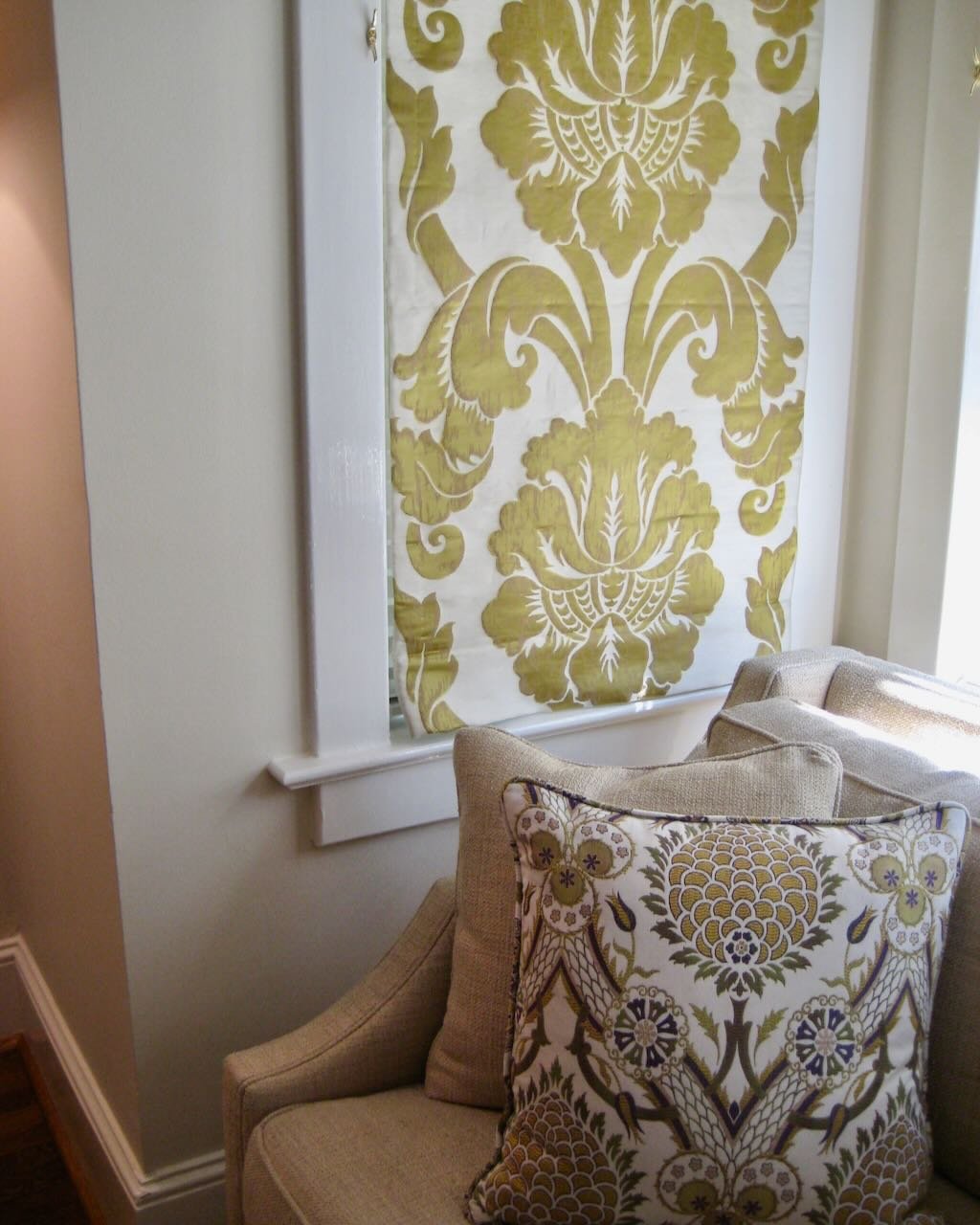 A window with a decorative, patterned yellow and white curtain, partially behind a beige armchair with patterned pillows.