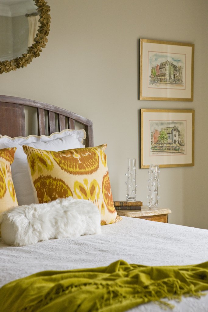 A cozy bedroom corner with a bed featuring a white cover and yellow patterned pillows, a small fluffy white rabbit, a wooden nightstand with glass candle holders, and framed watercolor art of buildings on the wall.