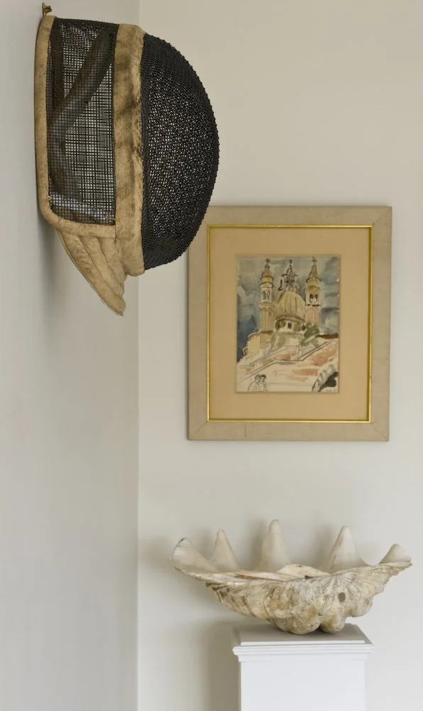 Decorative wall art and objects, including a framed painting of a church with domes, a large seashell, and a basket hat on a wall, with the hat hanging off a corner.
