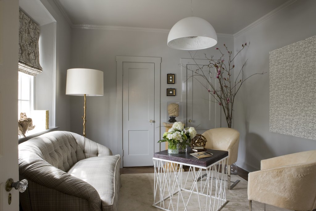 A cozy living room with a vintage-style sofa, a white leather armchair, a round dining table with floral decor, a tall floor lamp, window with Roman shade, and minimalist artwork on the wall.
