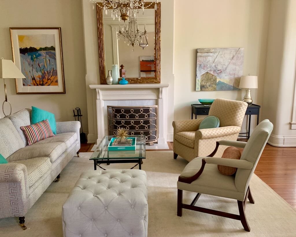 Living room with a white sofa, two armchairs, a glass coffee table, a white tufted ottoman, and wall art above the fireplace. A mirror and chandelier are reflected in the mirror above the fireplace. The room has neutral tones with colorful accents.
