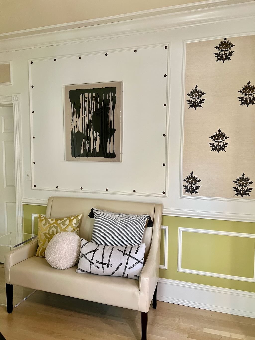 A living room wall with framed artwork, black and white patterned wallpaper on part of the wall, and a beige loveseat with patterned pillows in front of the wall.