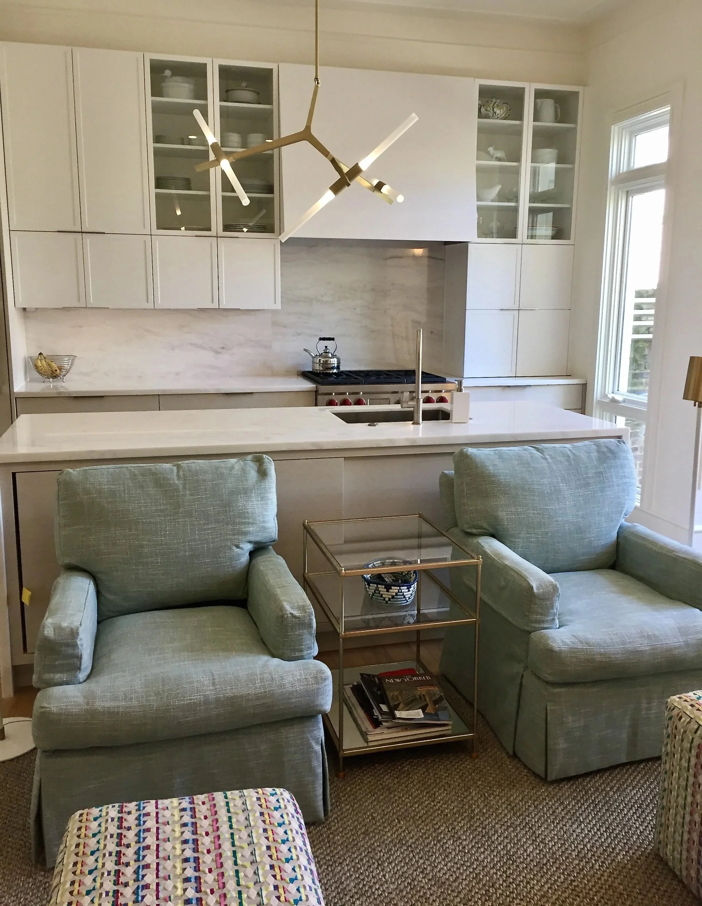 Living room with two light colored armchairs, a glass and gold side table with magazines or books underneath, and a modern kitchen with white cabinets, a marble countertop, and a gold geometric chandelier.
