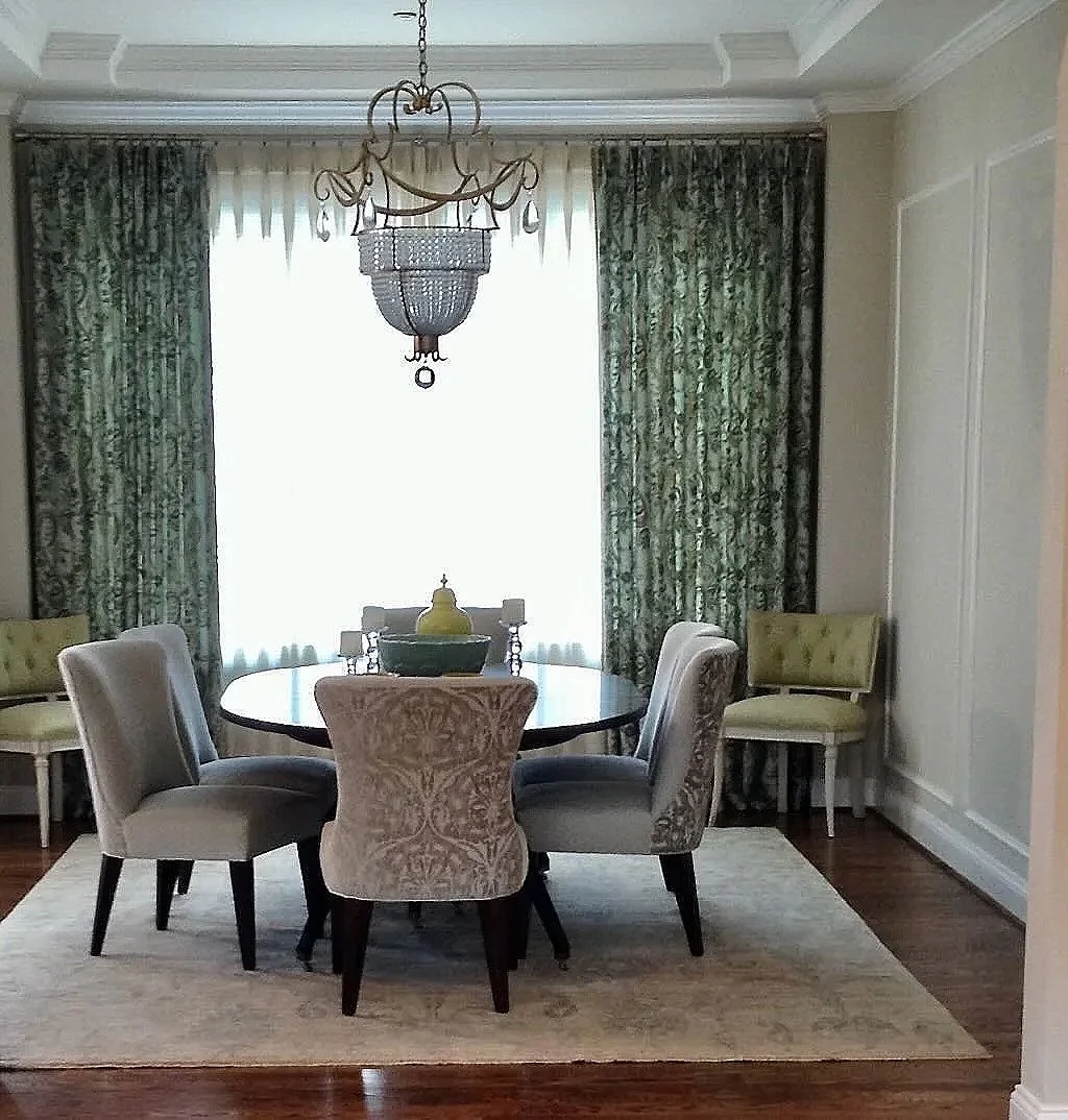 A dining room with a circular table surrounded by six upholstered chairs with varying patterns and colors, a chandelier hanging from the ceiling, dark green patterned curtains, and a light-colored rug on hardwood floor.