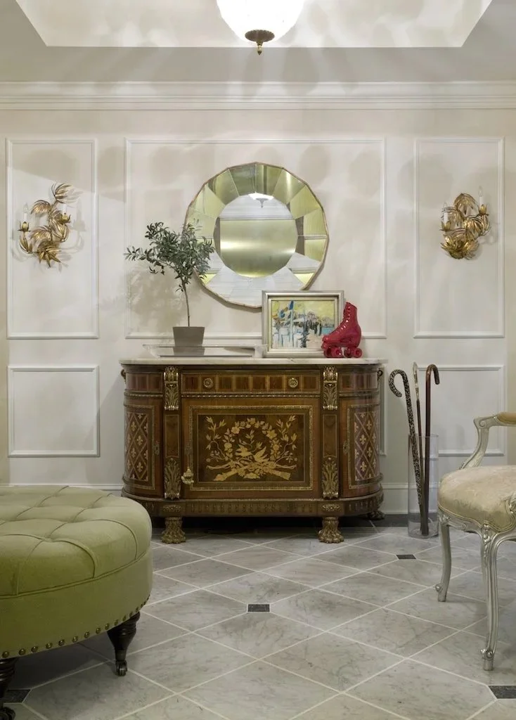 Elegant entryway with a wooden sideboard, gold wall sconces, a potted plant, a mirror, a framed painting, and a red roller skate sculpture on the sideboard. There is an olive green ottoman and a chair with light upholstery, and marble tile flooring.