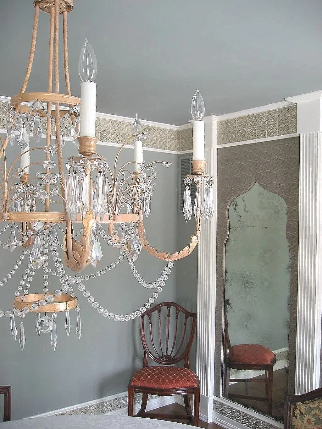A crystal chandelier with candle-shaped lights, hanging in a room with a vintage mirror, an antique chair with red upholstery, and decorative wallpaper.