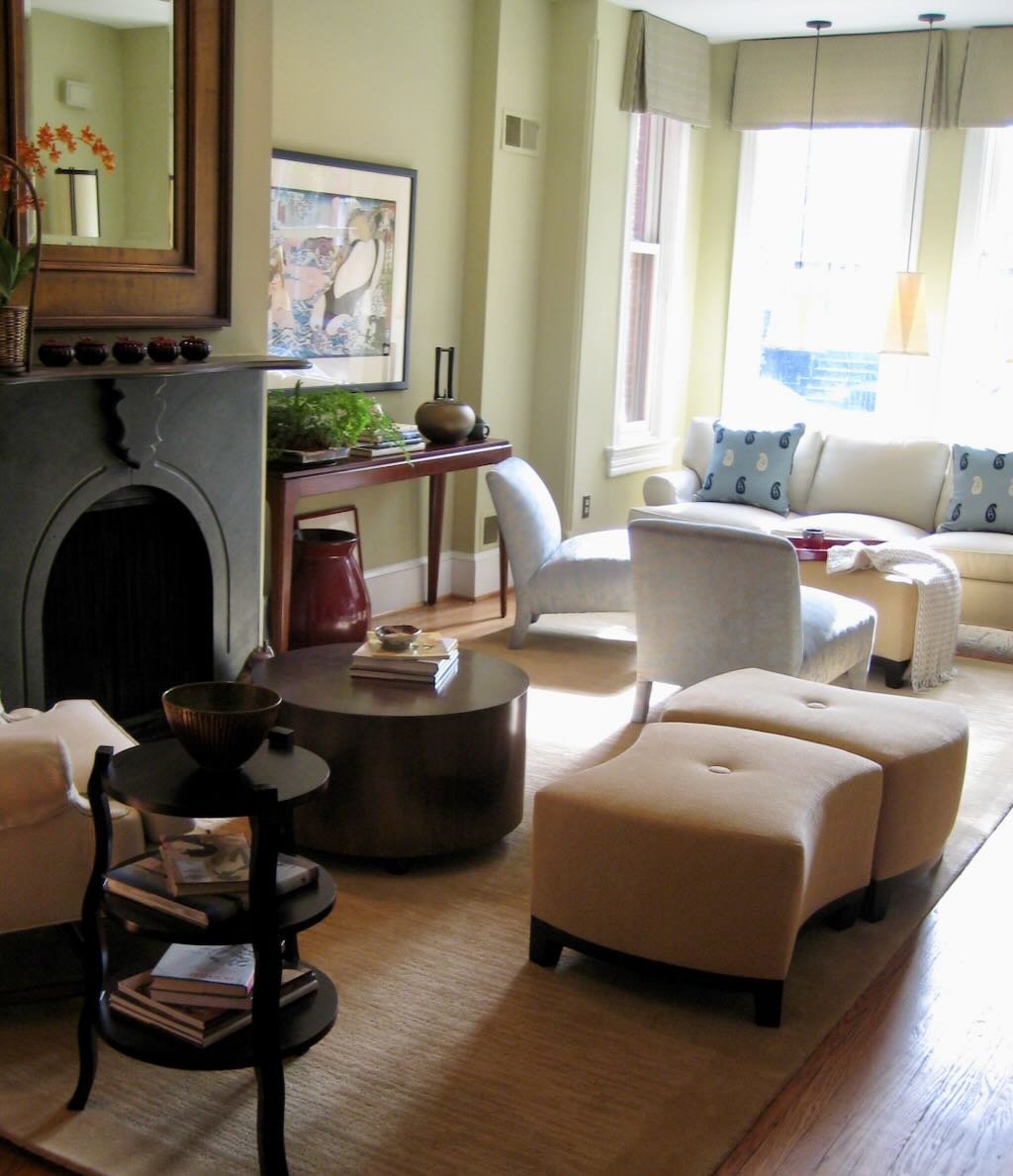 Living room with a fireplace, framed artwork, beige sofa with blue patterned pillows, beige armchair, ottoman, dark wood coffee table with books, and large windows with light green curtains allowing natural sunlight.