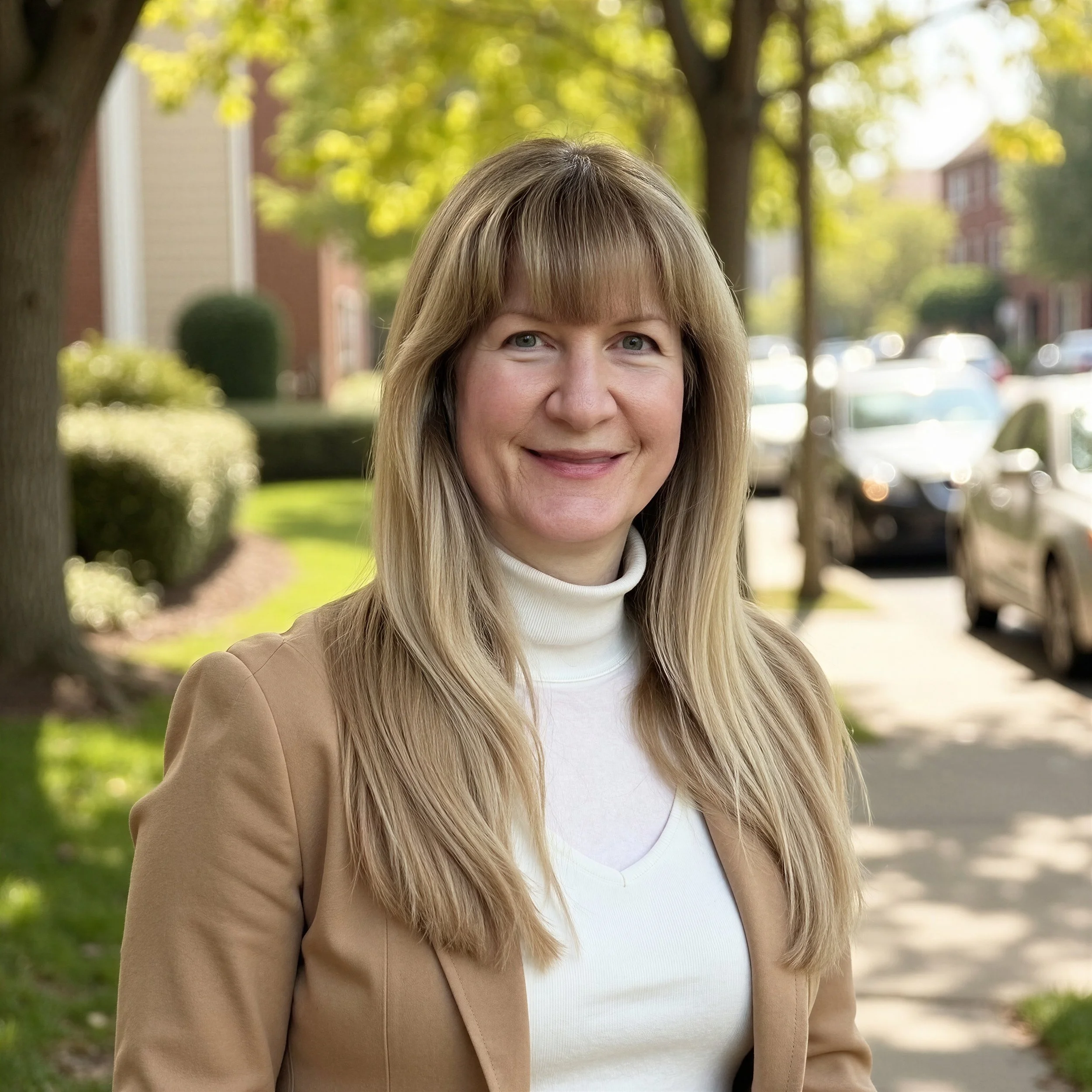Close-up portrait of a woman with blonde hair, blue eyes, and a friendly smile, outdoors on a sunny day with trees and parked cars in the background.