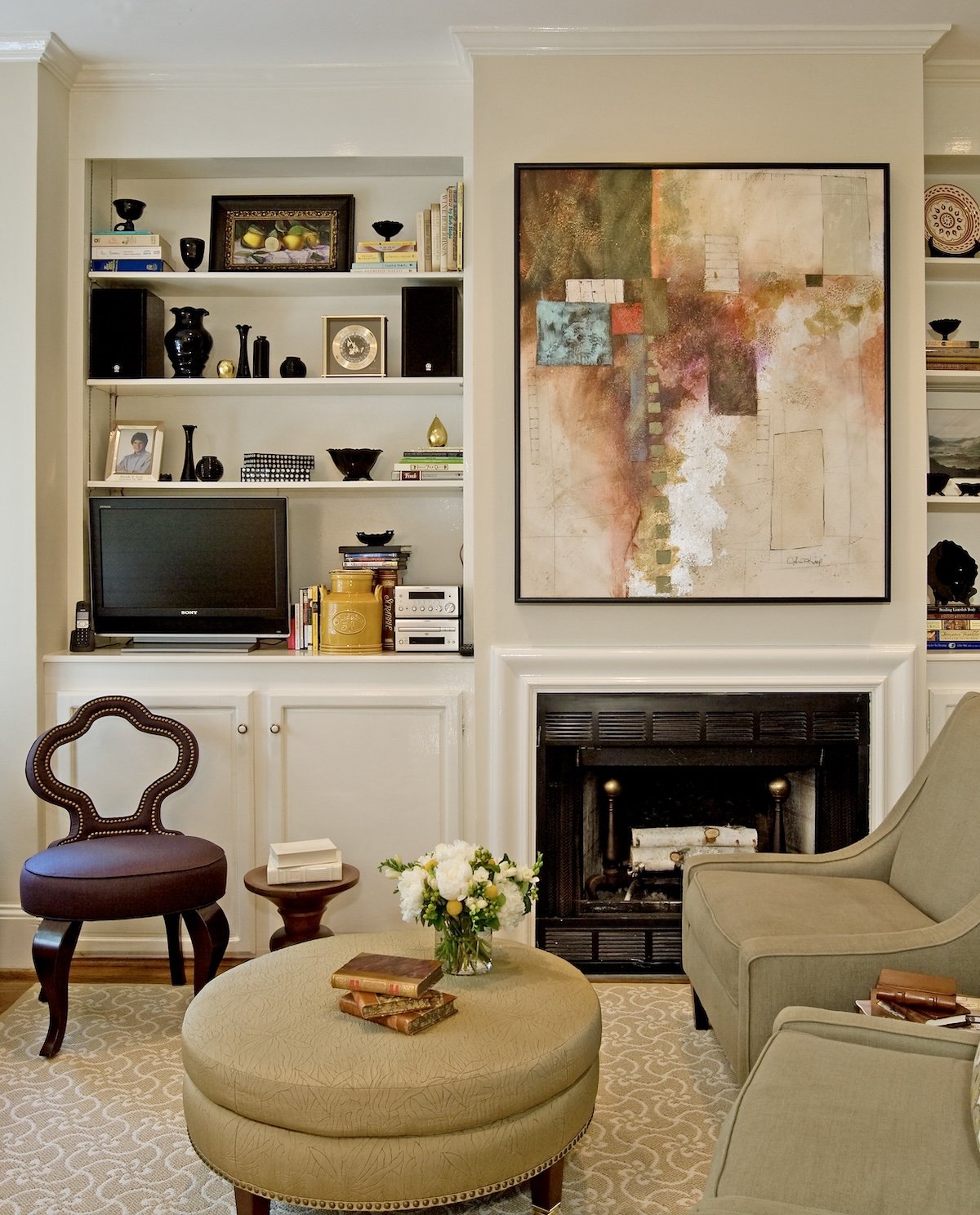 A living room with a white built-in bookshelf filled with books, decorative objects, and a small TV. There is a fireplace with a large abstract painting above, and a seating area with a beige ottoman, an armchair, and a side chair. A vase of white fl