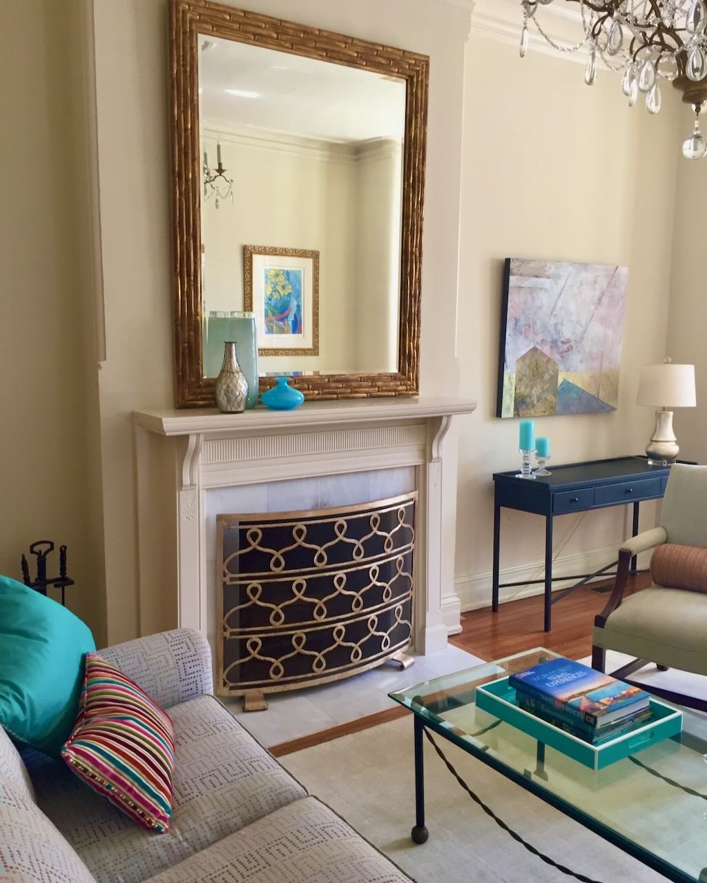 Living room with a large rectangular mirror above a white fireplace mantel, decorative vases on the mantel, a framed artwork on the wall, a black console table with blue candles, a beige armchair, a glass coffee table with books, a sofa with colorful