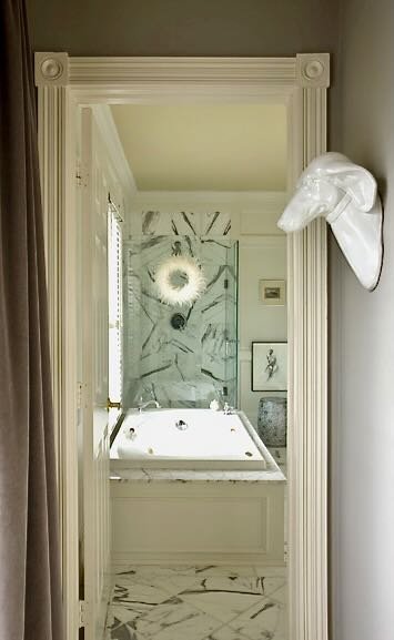 A bathroom with a white bathtub, decorative mirror, and art on the wall seen through a doorway. A white horse head sculpture is mounted on the right wall.