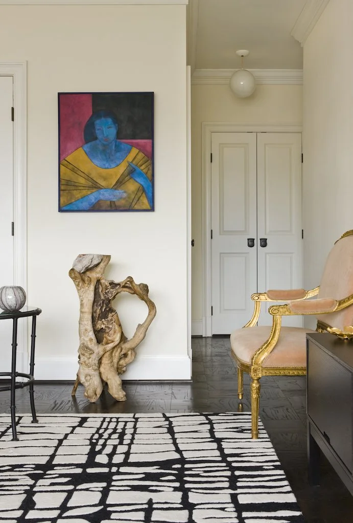 Interior of a room featuring a modern abstract painting of a woman, a rustic wooden sculpture, a vintage-style armchair with gold trim, a black side table with a decorative object, and a patterned black and white rug.