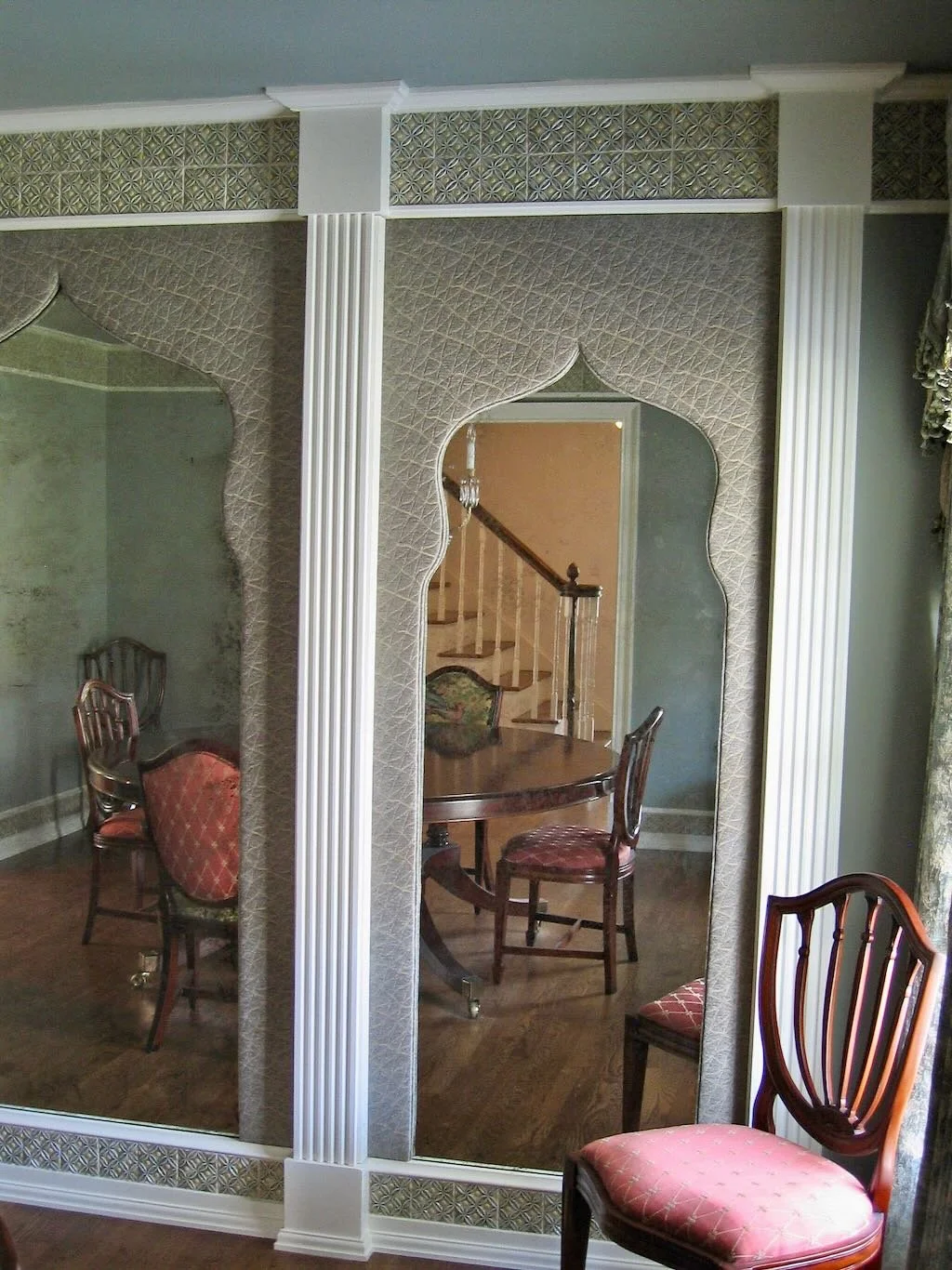 A decorated dining room featuring a large mirror with ornate framing, a round wooden dining table, and upholstered chairs with red patterned fabric. A staircase with a wooden banister is visible through the mirror's reflection.
