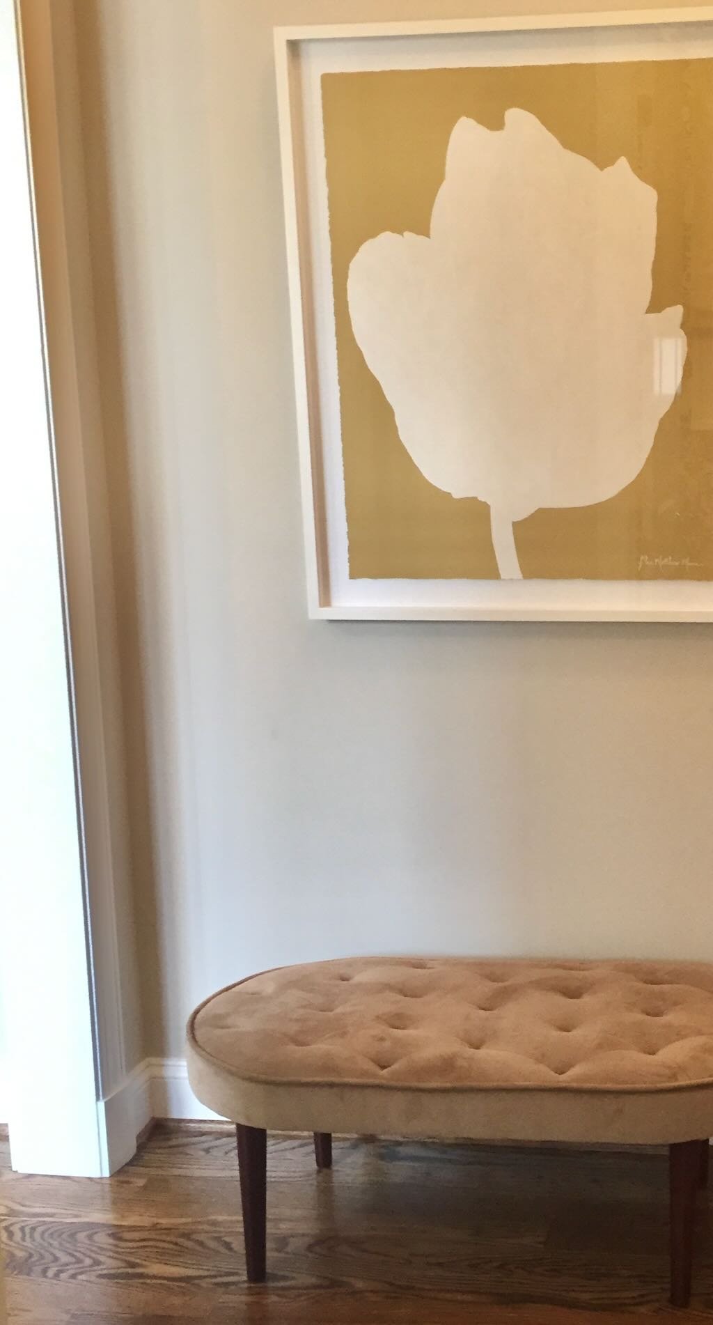 A beige upholstered bench with dark wooden legs placed on a wooden floor, next to a white wall with a framed artwork of a large white leaf on a gold background.