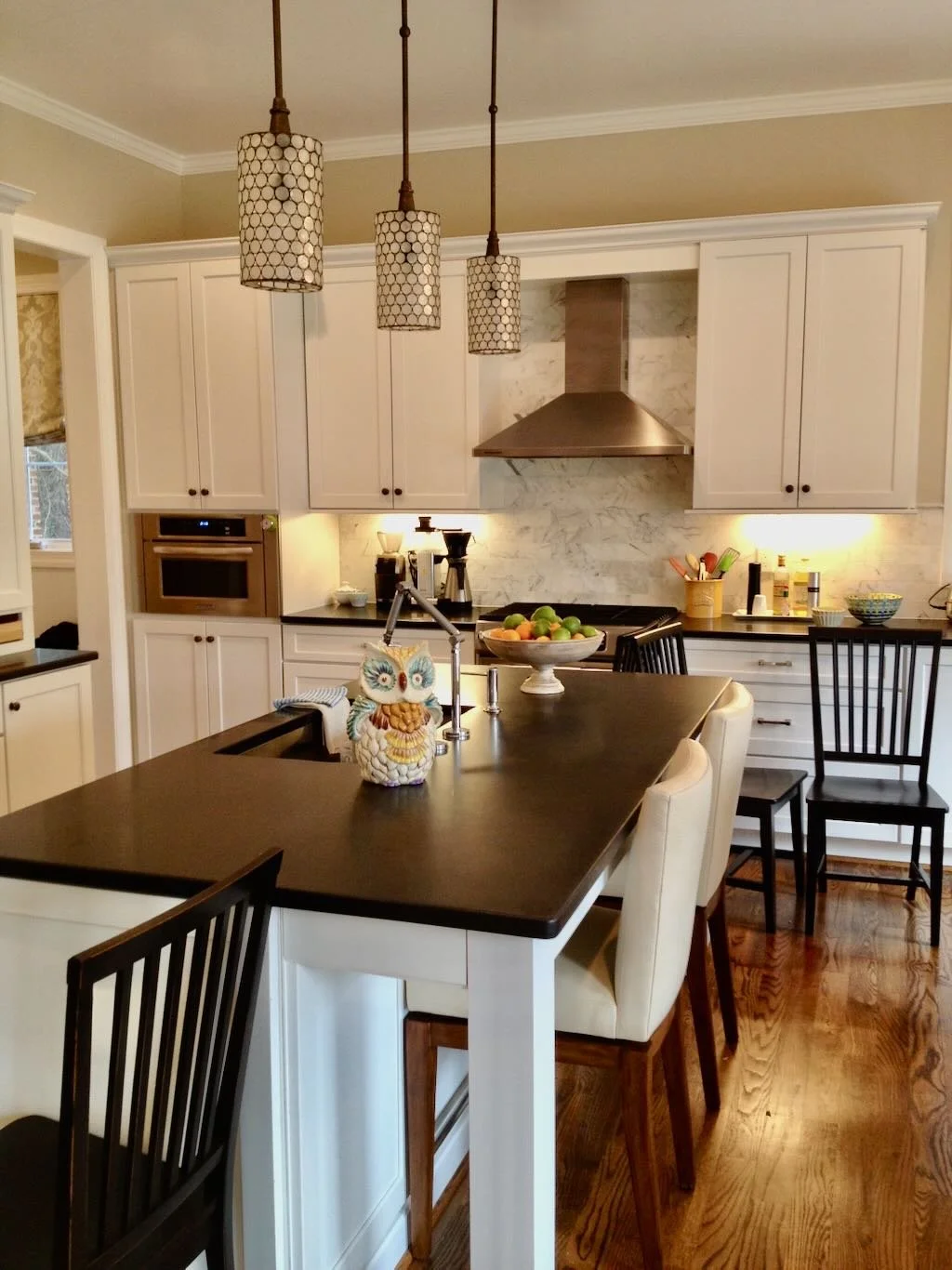 Modern kitchen with white cabinets, black countertops, and a large island. Above, three pendant lights hang over the island. There is a decorative owl figurine, a fruit bowl, and various kitchen items visible.