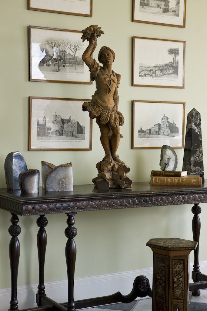 A vintage wooden table with a decorative edge and turned legs, featuring a classical sculpture of a young boy holding a hat, framed artwork of historical buildings, and various mineral or stone specimens. A small ornate wooden stool is positioned in 