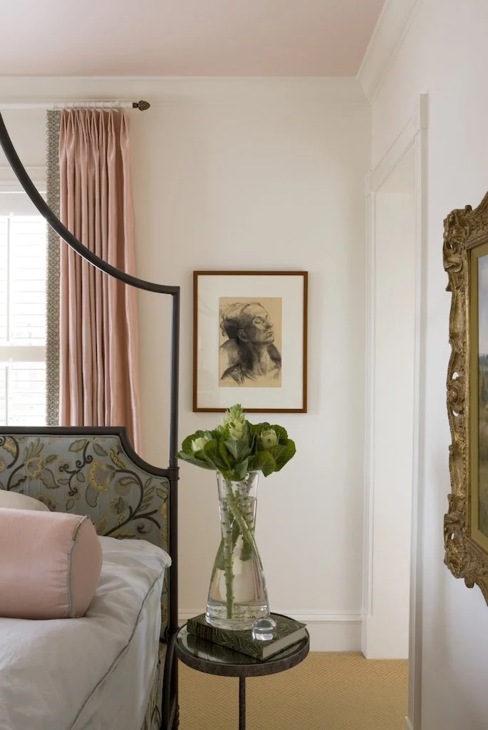 A bedroom with a bed, a nightstand with a vase of green flowers, pink curtains, a framed portrait on the wall, and part of an ornate gold frame mirror.