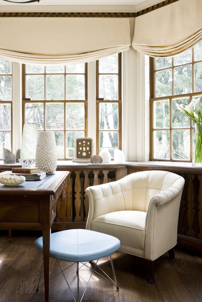 A cozy corner of a room with large multi-pane windows, decorated with beige curtains, featuring vintage furniture including a cream leather armchair and a white modern ottoman, a wooden desk, and decorative vases and objects on the window ledge.