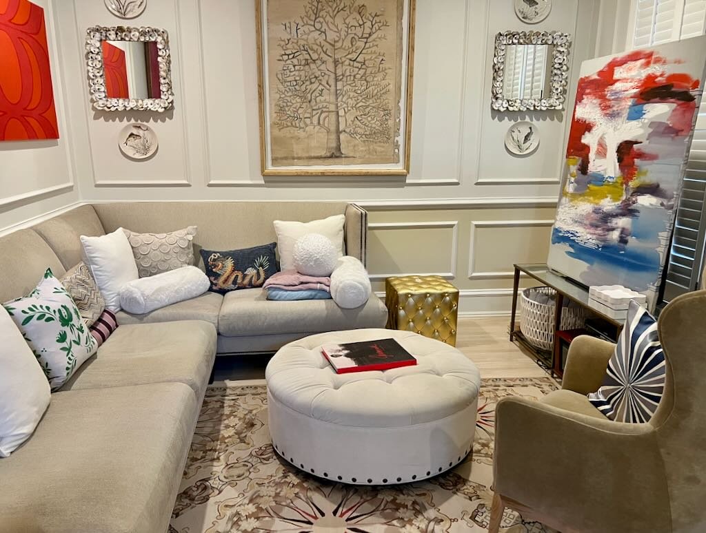Living room with beige sectional sofa, decorative pillows, framed artwork, mirrors, and an abstract painting on an easel.