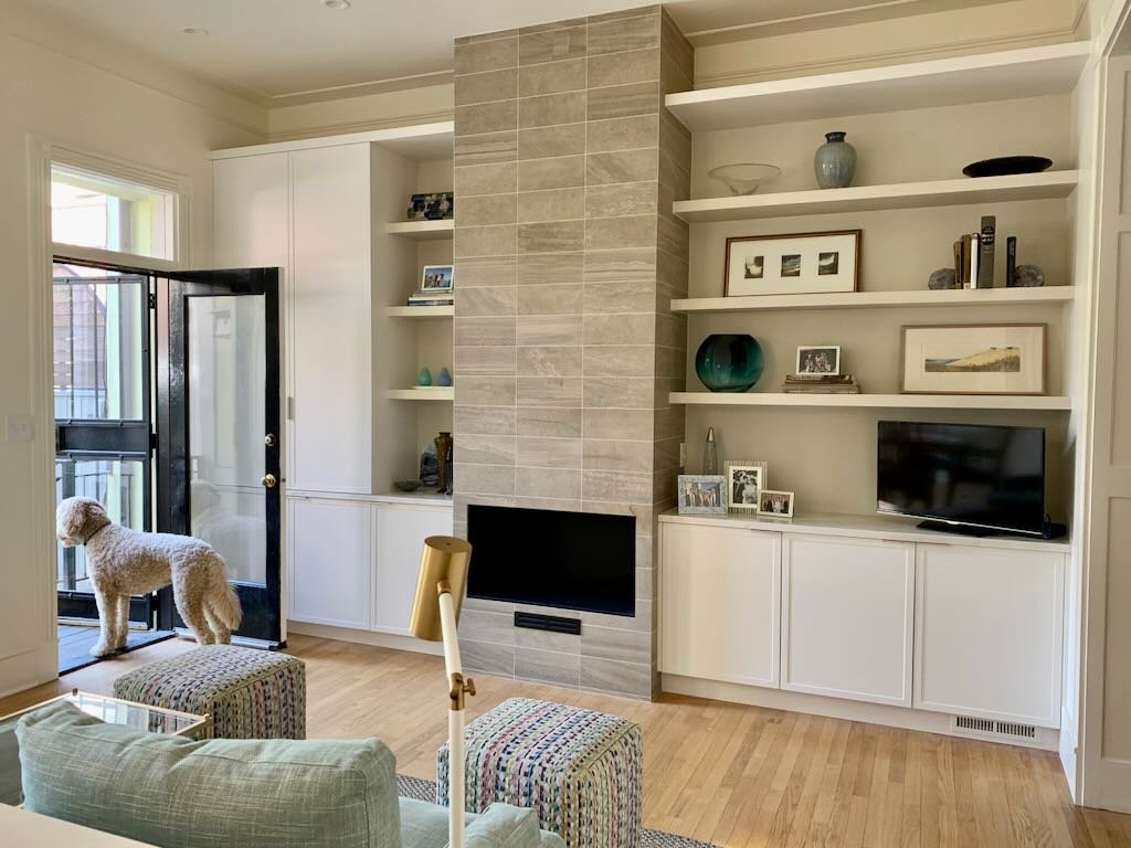 Living room with a fireplace, built-in white shelves with decorative items and framed pictures, a small TV, and a large window/door with a dog looking outside.