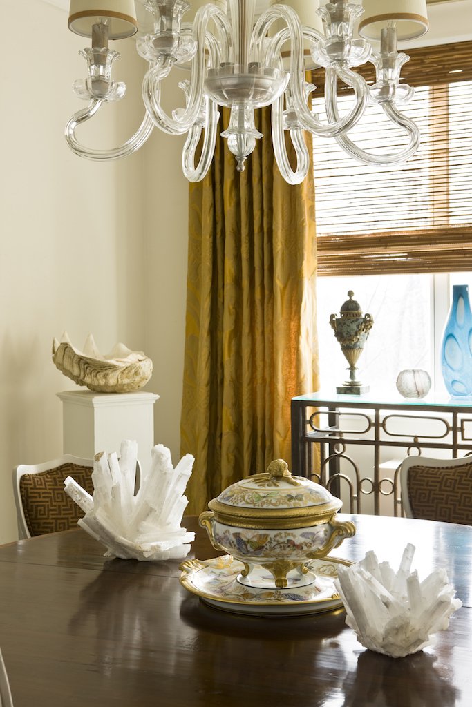 A chandelier with glass arms hanging above a dining table, which is decorated with ornate dishes and crystal-like sculptures. The background features a window with wooden blinds and curtains, as well as shelves with decorative vases and artifacts.