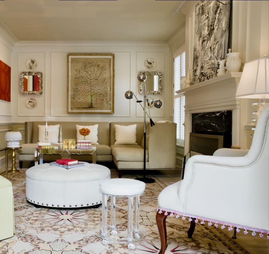 A living room with beige and white furniture, including a couch, armchair, ottoman, and small table, with decorative art on the walls, a fireplace, and a patterned rug.