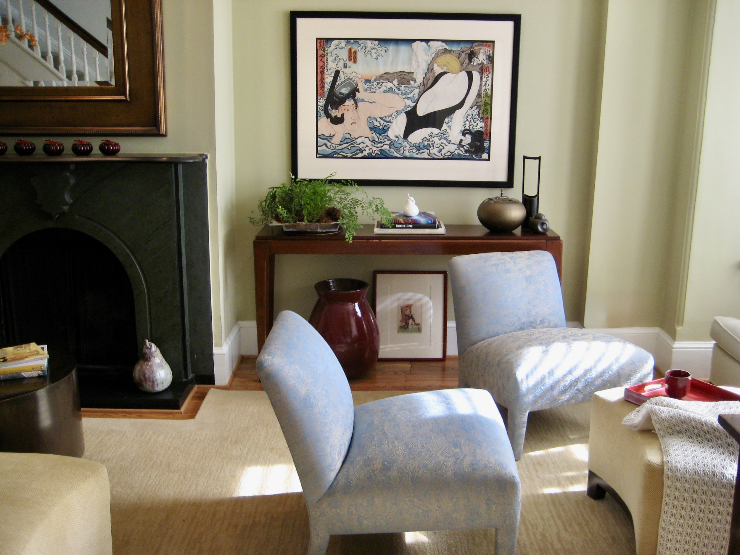 A cozy living room corner with two light blue upholstered chairs, a beige sofa, a fireplace, a wooden console table with decorative items, and a large framed Japanese-style artwork on the wall.