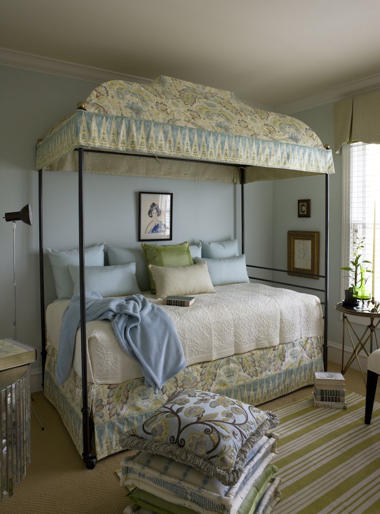 A bedroom with a canopy bed featuring a floral pattern and an ornate headboard, multiple pillows, a blanket, a picture hanging on the wall, and a window with blinds.