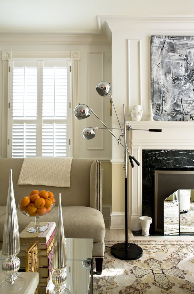 A contemporary living room with a beige sofa, a glass coffee table, metallic vases, and a modern floor lamp with three spherical shades. There's a fireplace with a black marble surround, artwork above, and a window with white shutters.