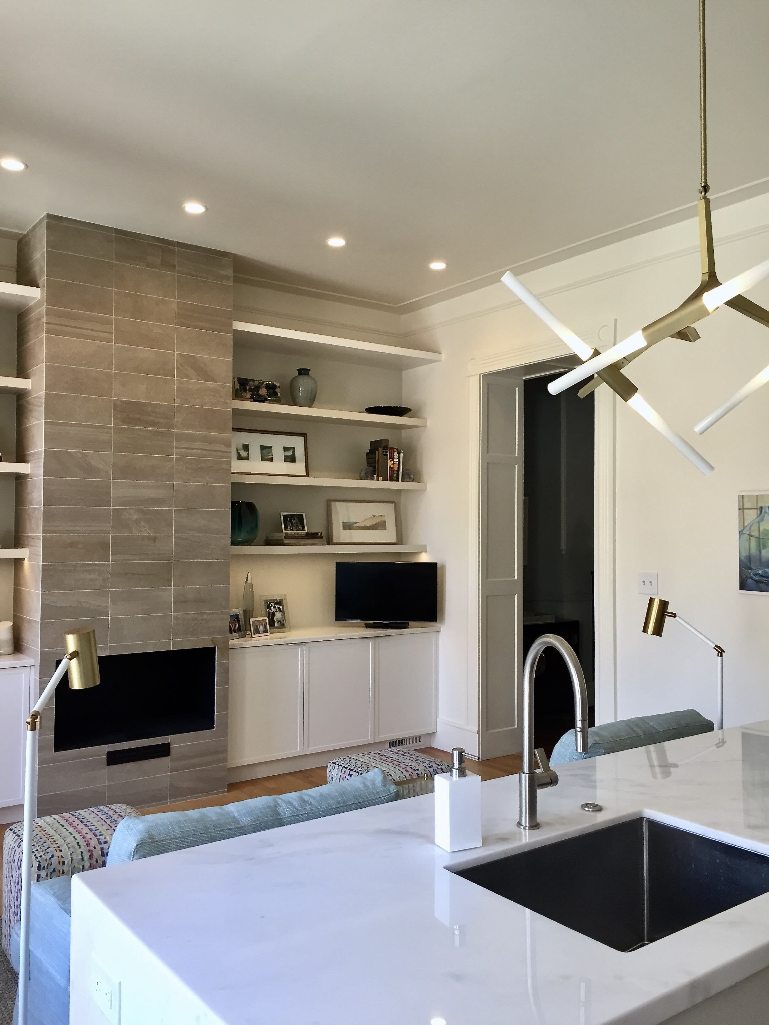 Modern kitchen with white countertops, gold and white lighting fixtures, built-in white shelving with decor and a TV, beige tiled fireplace, and a doorway leading to another room.