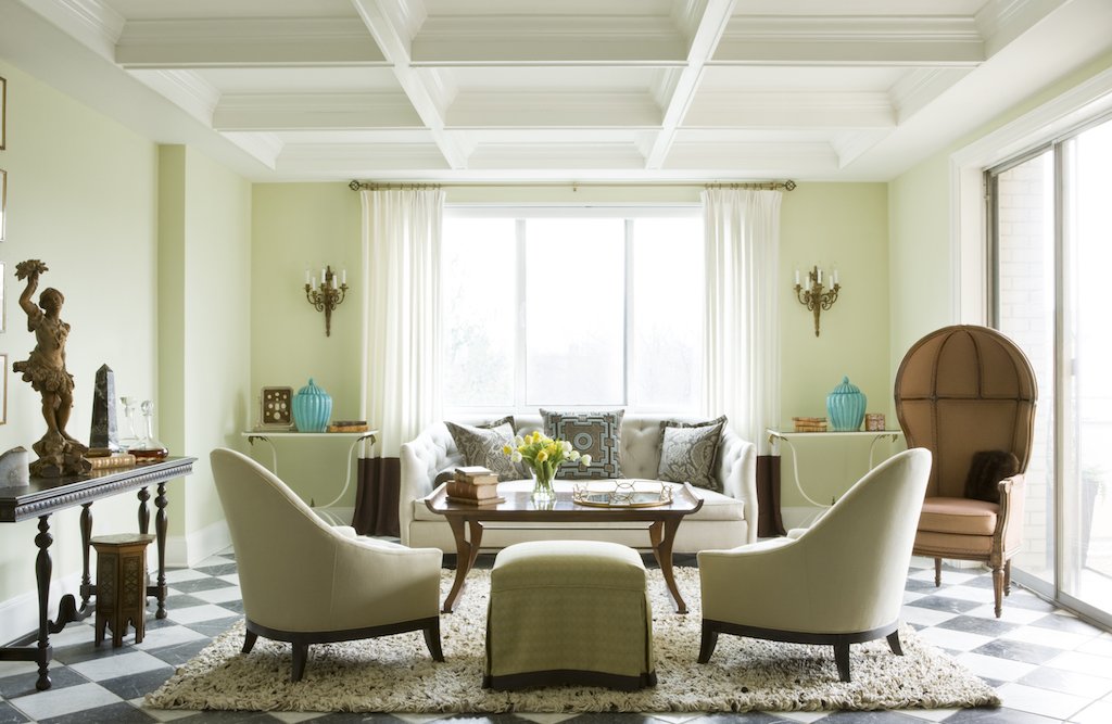 Bright living room with light green walls, large window with white curtains, white sofa with decorative pillows, two white armchairs, a beige wingback chair, a wooden coffee table with books and a flower centerpiece, black-and-white checkered floor, 