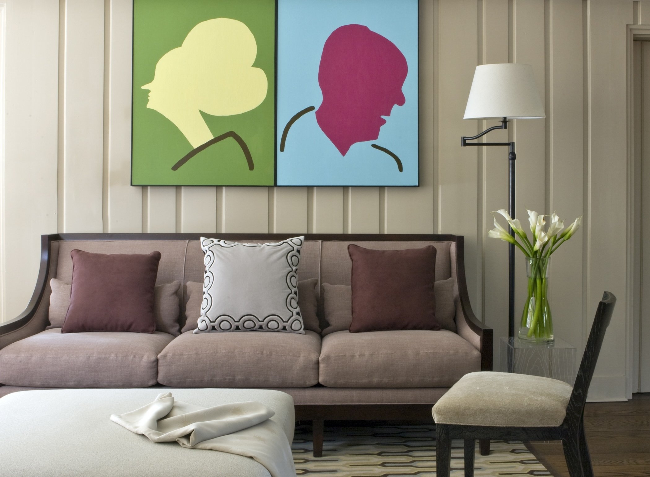 Living room with a beige sofa, purple and off-white pillows, a glass table with white cloth, a black chair, a tall black floor lamp, and a vase of white lilies. There are two colorful abstract portraits of faces hanging on the wall.