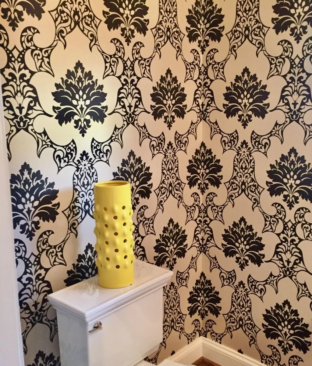 A yellow ceramic vase with holes placed on top of a white cabinet in front of a black and white damask patterned wallpaper.
