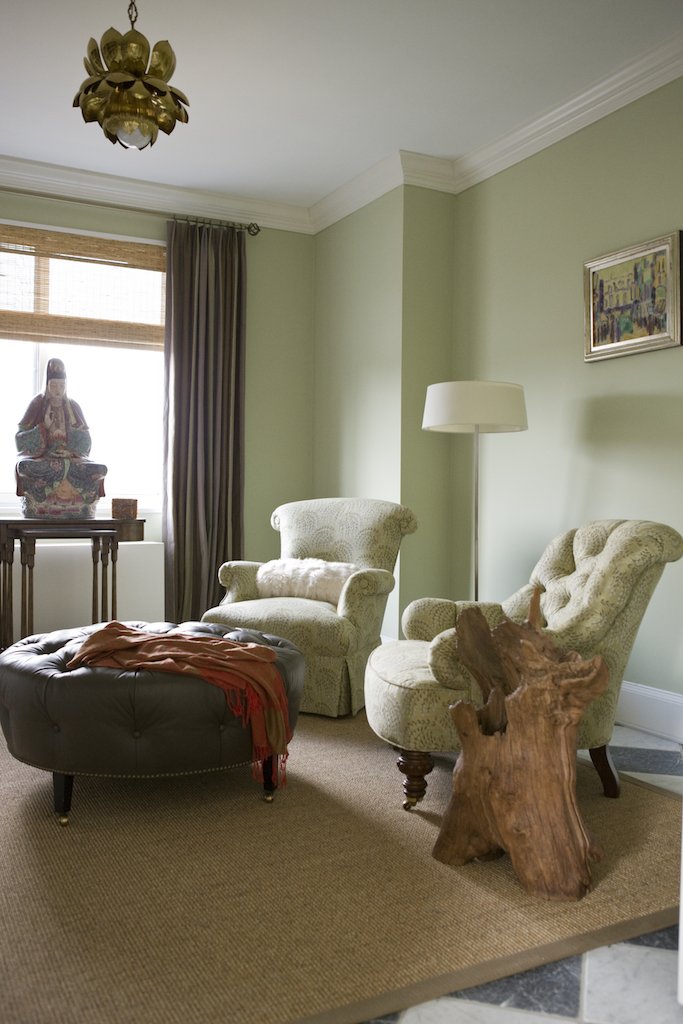 Living room with green walls, two beige armchairs, a black ottoman with a scarf, a wooden side table with a Buddha statue, and a floor lamp. Window with curtains and a framed painting on the wall.