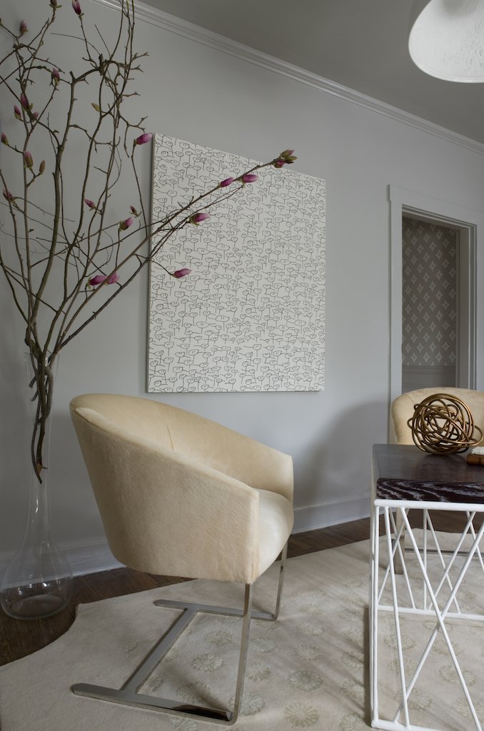 A cozy living room corner with a cream upholstered armchair, a large glass vase holding blooming branches, an abstract black and white artwork on the wall, and a small gray table with a decorative woven ball on top.