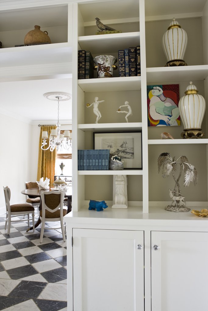 View of a white built-in bookshelf with decorative items, including sculptures, vases, and books, in a dining area with checkered black-and-white tiled floor and a dining table with chairs.