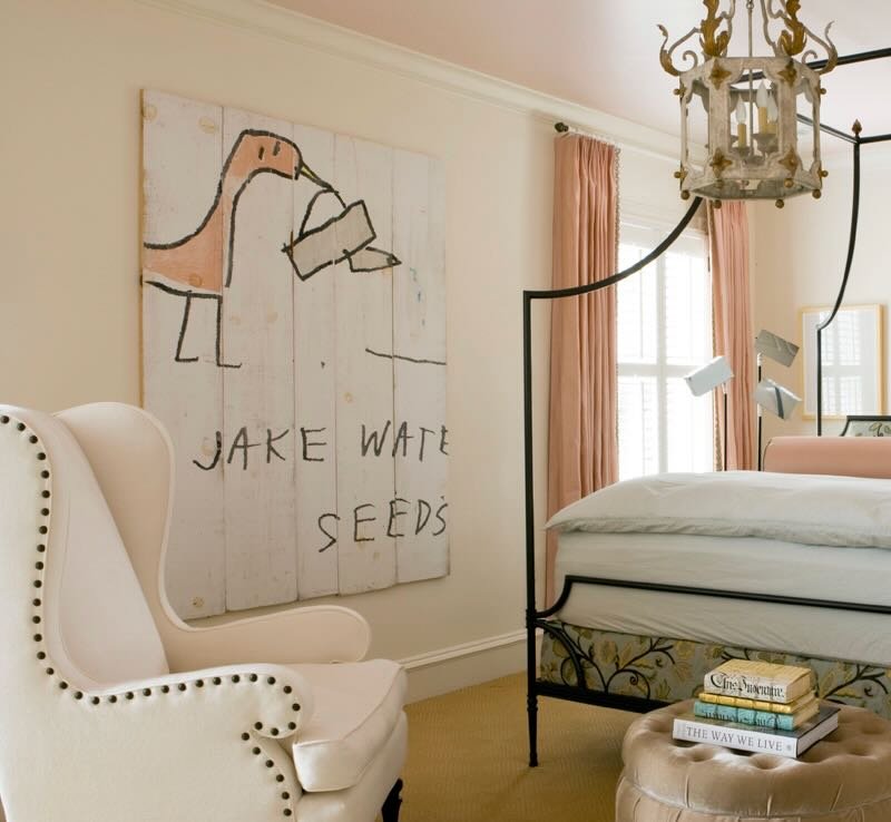 A cozy bedroom with a white wingback chair, a large framed wall art with a child's drawing of a hand watering seeds, pink curtains, a black metal bed frame, a small stack of books on a tufted ottoman, and a vintage chandelier hanging from the ceiling