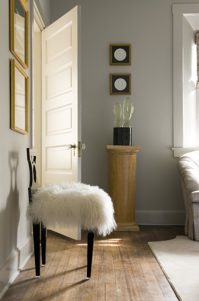 Interior living space with a white upholstered chair with black legs and fluffy fur cover, a wall with framed artwork, a wooden pedestal with decorative vases, a partially open door, and a window with sunlight, hardwood floor, and part of a sofa.