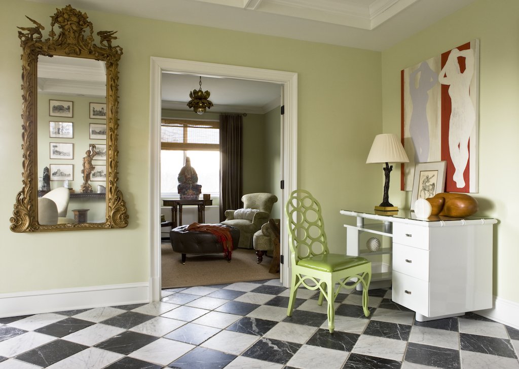 A view into a living room through an open doorway, showcasing eclectic decor including a vintage mirror, a green chair, a white desk with decorative items, and framed artwork on the wall, with a checkerboard black-and-white tiled floor.