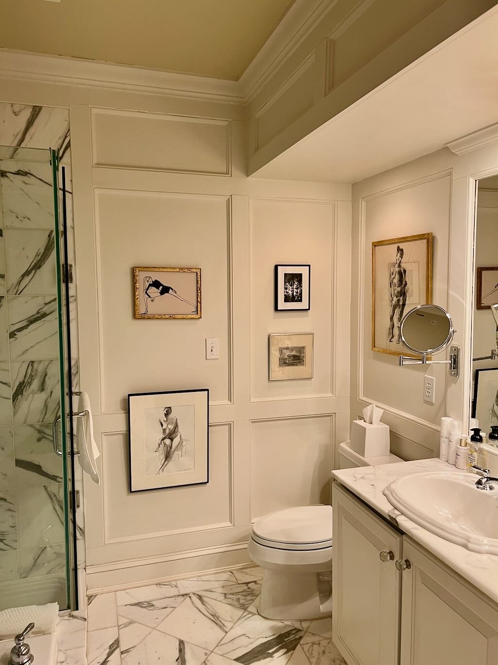 A bathroom with white walls decorated with black and white art, a marble floor, a shower stall with marble tiling, a white vanity with a marble top, a round mirror, tissue box, and personal care products.