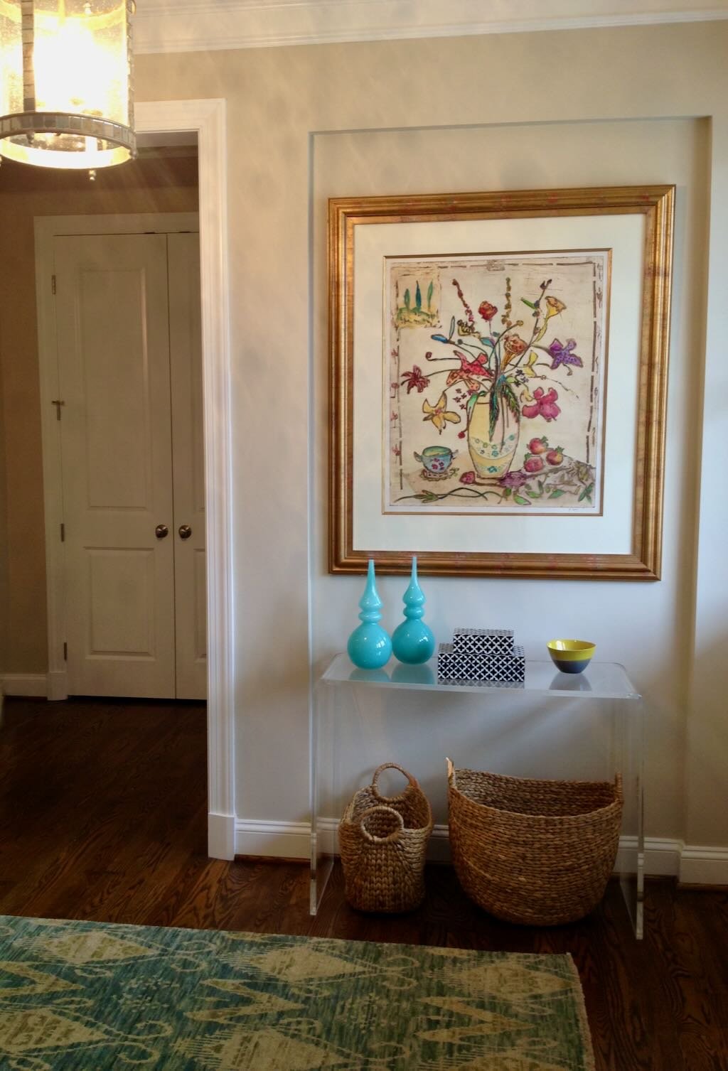 A hallway corner with a decorative framed artwork of colorful flowers in a vase above a white console table. The table holds two turquoise vases, a black and white patterned box, and a black and yellow bowl. Below the table are two woven baskets, one