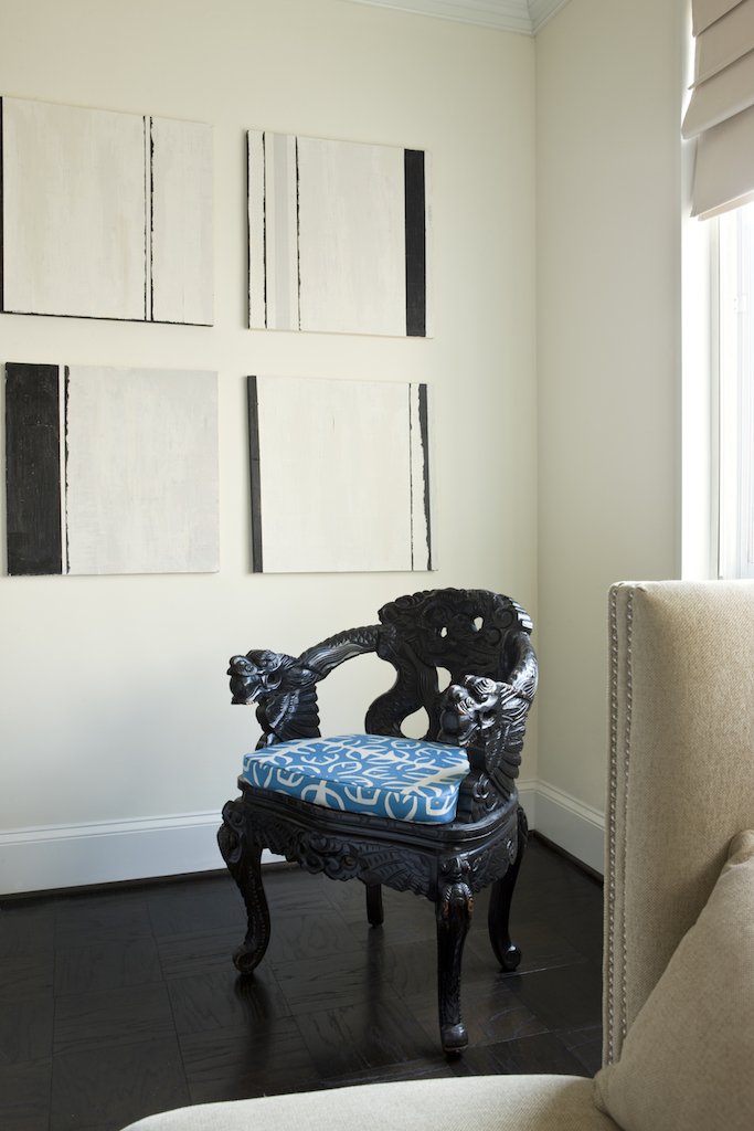 A black carved antique chair with a blue and white patterned cushion, placed in a corner of a room with white walls, a beige upholstered chair to the right, and abstract black and white artwork on the wall.
