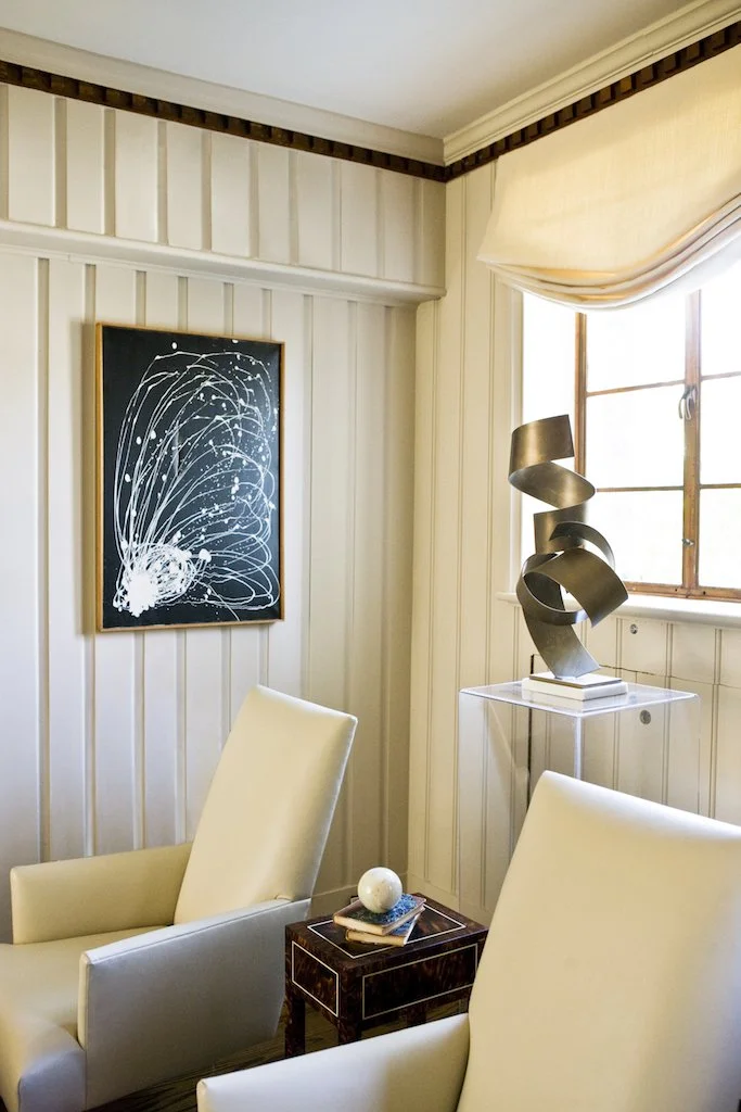 A cozy corner of a room with two cream-colored armchairs, a small dark wood side table with a book and decorative orb, a black abstract art piece on the wall, a modern sculpture on a clear table, and a window with beige drapes allowing natural light 