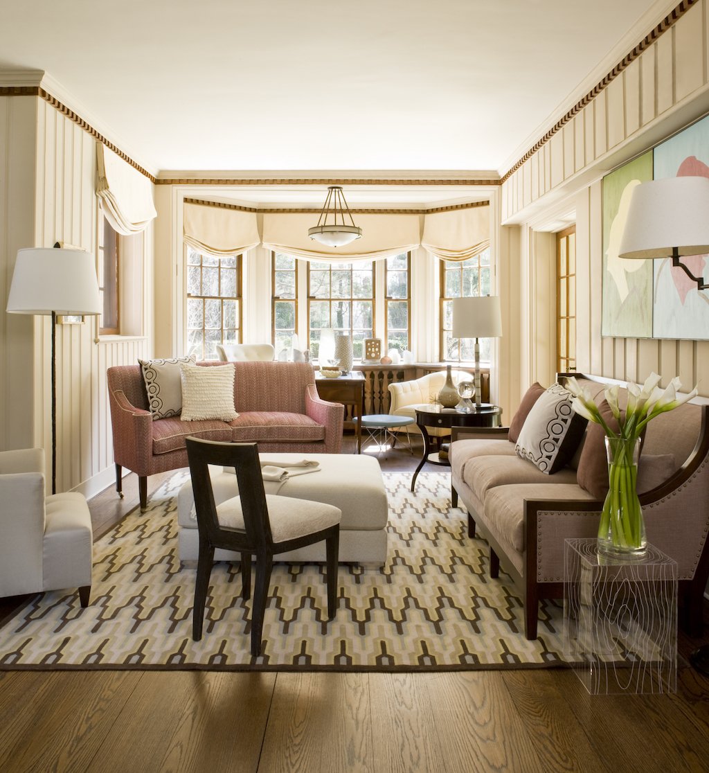 A cozy living room with wooden floors, cream-colored paneled walls, and large bay windows with cream valances. The room features a pink sofa, beige sofa, armchair, and various side tables with lamps. A patterned rug covers the floor, and a vase of wh